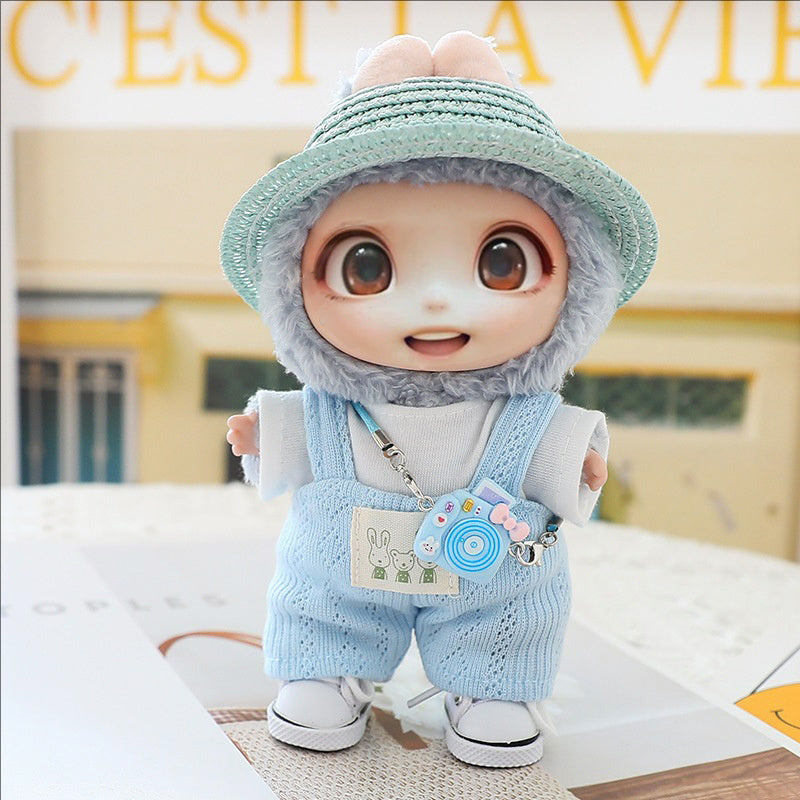 Cute doll wearing blue knitted hat, matching blue overalls, white shoes, and carrying a small toy camera accessory