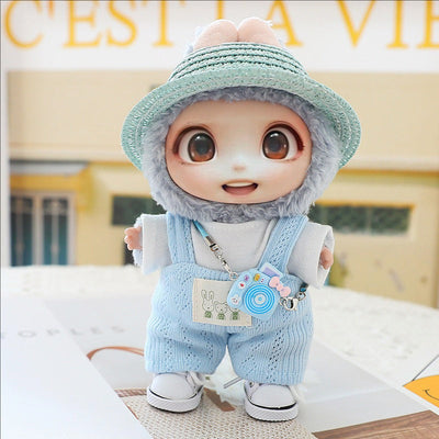 Cute doll wearing blue knitted hat, matching blue overalls, white shoes, and carrying a small toy camera accessory
